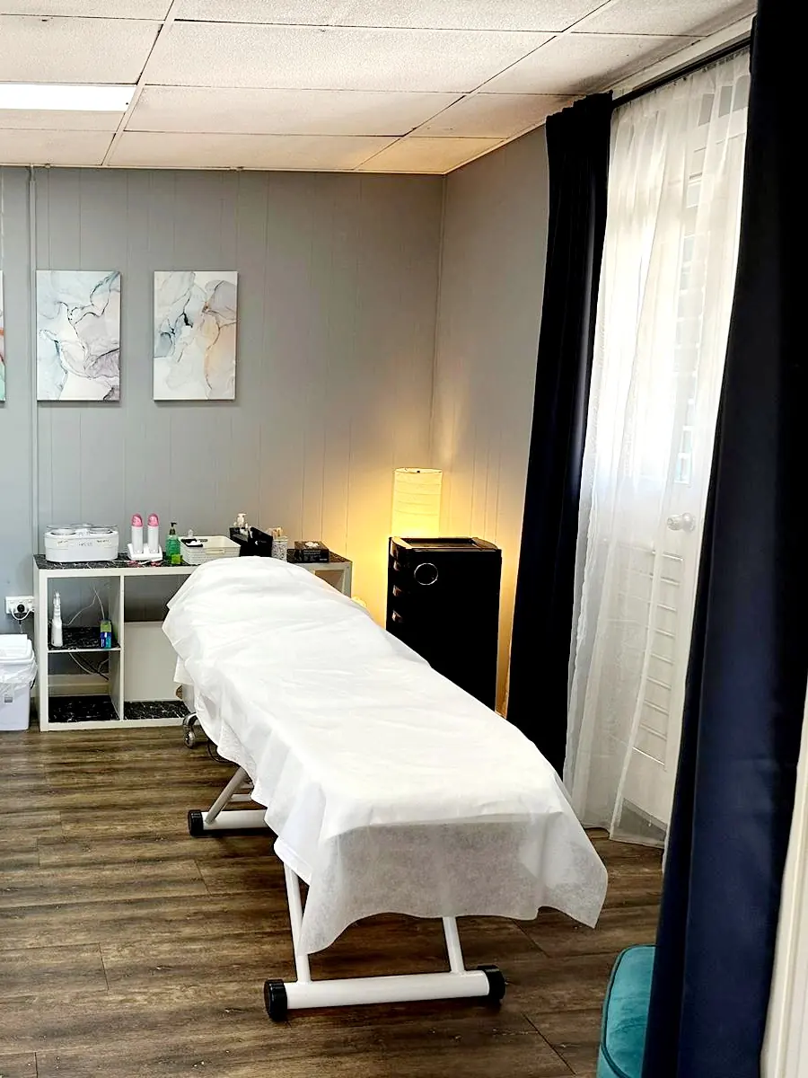 A white-draped treatment bed beside a warm lamp, with wax heaters and products on the marble shelf